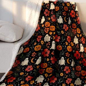 May include: A black Halloween-themed blanket with a pattern of orange pumpkins, white ghosts, red flowers, and crescent moons. The blanket has a soft, white sherpa lining on the underside and is draped over a beige sofa.