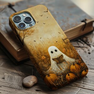 May include: A smartphone case featuring a printed illustration of a white ghost reading a book in a golden autumn field with orange pumpkins, tall dry grass, and small black bats flying in the sky against a warm, sepia-toned background.