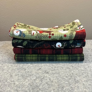 May include: A stack of four folded fabric squares with holiday-themed patterns. The top square features snowmen on a green background. Below are squares with holly, red plaid, and green plaid designs. The fabrics are stacked on a gray surface.