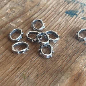 May include: A group of eight silver metal rings with a textured surface and small raised dots around the edges. The rings are arranged in a loose pile on a wooden surface.