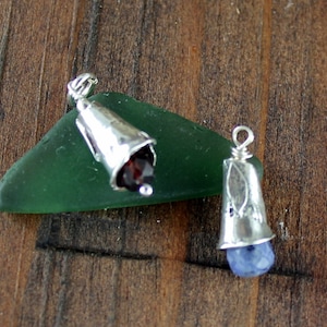 May include: Two silver pendant charms with gemstone accents. The charm on the left has a red gemstone and the charm on the right has a blue gemstone.