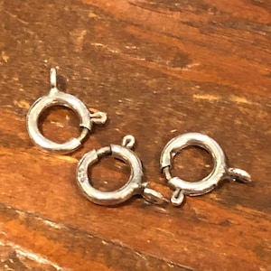 Puede incluir: Tres pequeños cierres de anillo de resorte plateados. Cada cierre es circular con un pequeño lazo y un mecanismo de resorte para un cierre seguro. Los cierres están sobre una superficie de madera.