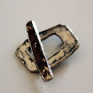 May include: A silver pendant with a rectangular shape and a smaller rectangular cutout in the center. The pendant has a textured surface and a silver bar that crosses over the top.