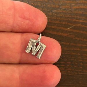 May include: A small, silver-colored pendant in the shape of the letter "M". The pendant has a textured, hammered appearance and a small loop at the top for a chain. The pendant is held between two fingers.