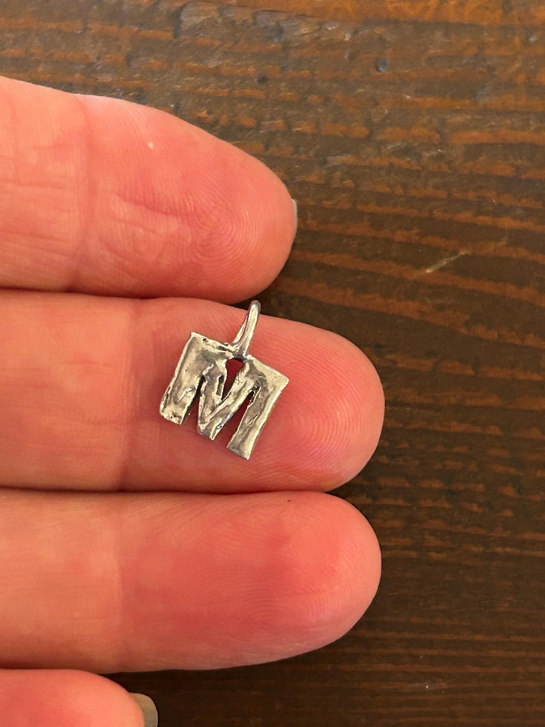 May include: A small, silver-colored pendant in the shape of the letter "M". The pendant has a textured, hammered appearance and a small loop at the top for a chain. The pendant is held between two fingers.