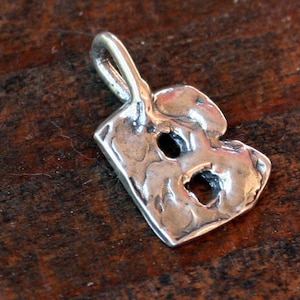 May include: A silver-colored pendant in the shape of the letter "B". The pendant has a textured surface with two holes and a loop at the top for a chain. The pendant is displayed on a dark wooden surface.