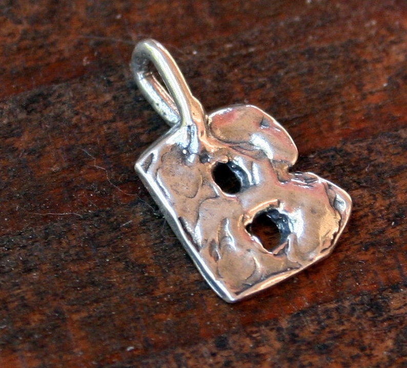 May include: A silver-colored pendant in the shape of the letter "B". The pendant has a textured surface with two holes and a loop at the top for a chain. The pendant is displayed on a dark wooden surface.