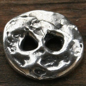 May include: A silver charm in the shape of a skull with two eye holes. The charm has a rough, textured surface.