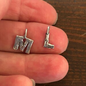 May include: Two small, silver-colored metal charms in the shape of the letters "M" and "L". The charms have a textured, handmade appearance and small loops at the top for attaching to a chain or bracelet. The "M" charm is larger than the "L" charm.