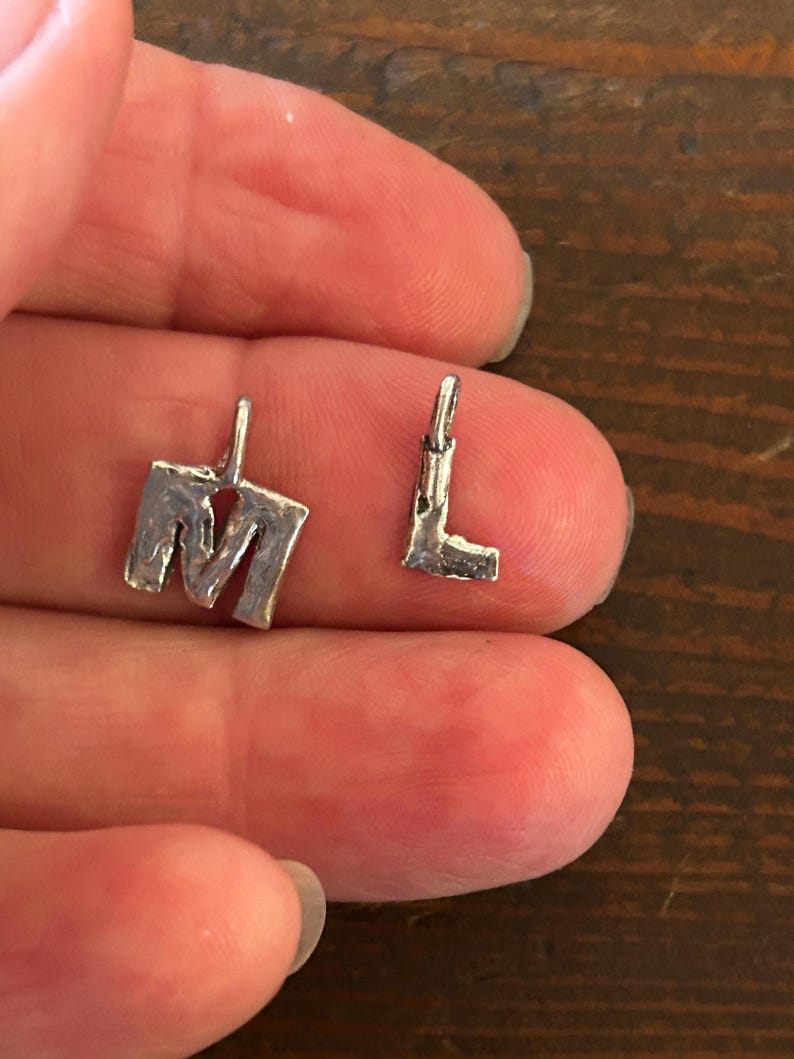 May include: Two small, silver-colored metal charms in the shape of the letters "M" and "L". The charms have a textured, handmade appearance and small loops at the top for attaching to a chain or bracelet. The "M" charm is larger than the "L" charm.