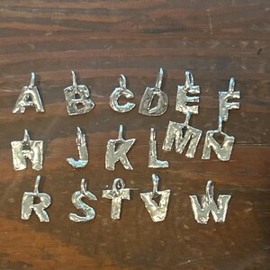 May include: Silver-toned alphabet pendants, each with a unique, textured design. The letters A, B, C, D, E, F, H, J, K, L, M, N, R, S, T, and W are displayed. The pendants are suspended from small loops.