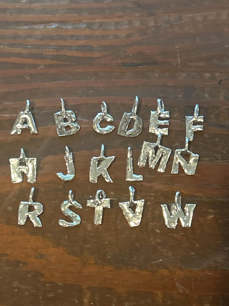 May include: Silver-toned alphabet pendants, each with a unique, textured design. The letters A, B, C, D, E, F, H, J, K, L, M, N, R, S, T, and W are displayed. The pendants are suspended from small loops.