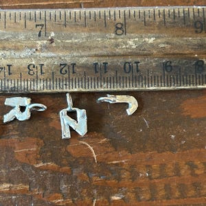 May include: Close-up of three small silver metal charms, including the letters "R" and "N", and a hook shape. A wooden ruler with inch measurements is in the background. The charms are on a wooden surface.