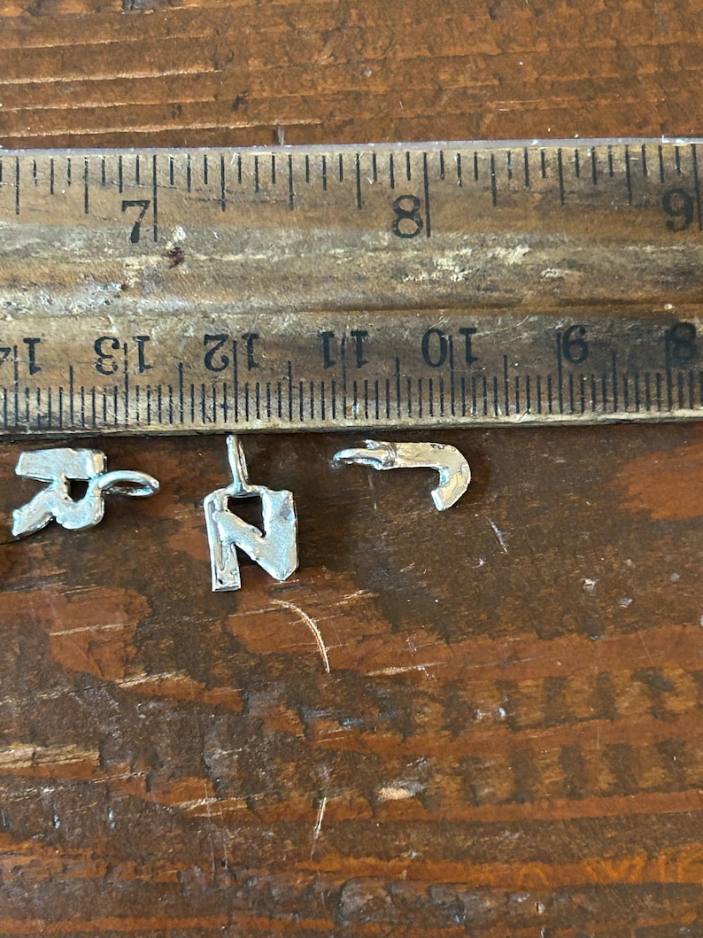 May include: Close-up of three small silver metal charms, including the letters "R" and "N", and a hook shape. A wooden ruler with inch measurements is in the background. The charms are on a wooden surface.