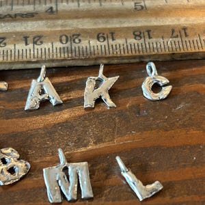 May include: Close-up of several small, silver-colored metal letter charms. The letters 'A', 'K', 'C', 'M', and 'L' are visible. A wooden ruler with inch markings is in the background.