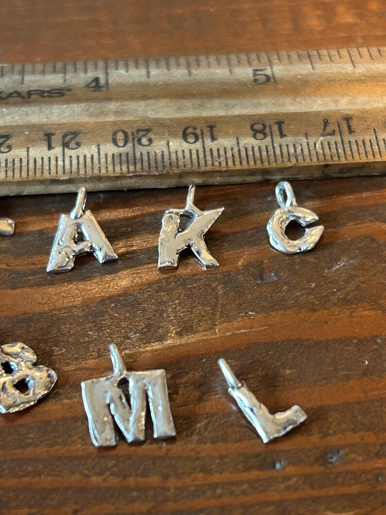 May include: Close-up of several small, silver-colored metal letter charms. The letters 'A', 'K', 'C', 'M', and 'L' are visible. A wooden ruler with inch markings is in the background.