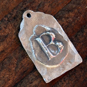 May include: A silver charm with the letter "B" embossed on it. The charm is shaped like a tag and has a hole at the top for hanging.