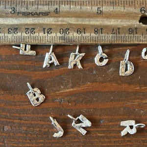 May include: A collection of small, silver-colored alphabet charms, each with a loop for attaching to a chain or bracelet. The charms are arranged on a wooden surface next to a ruler, showing their size.