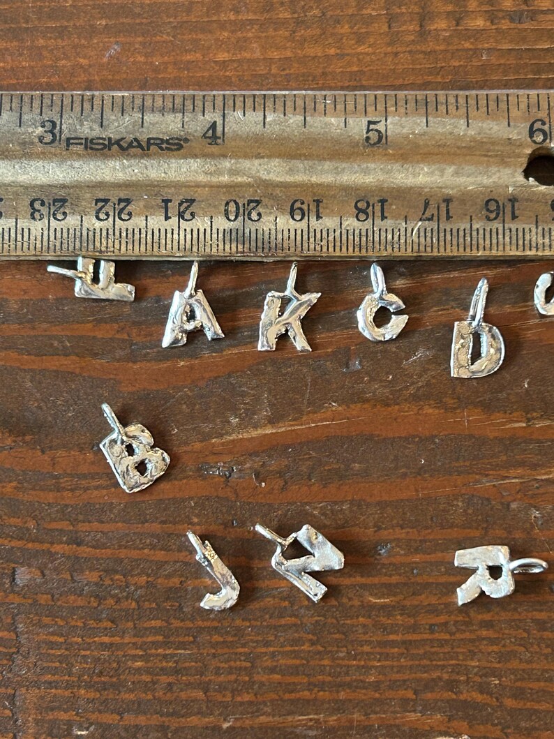 May include: A collection of small, silver-colored alphabet charms, each with a loop for attaching to a chain or bracelet. The charms are arranged on a wooden surface next to a ruler, showing their size.