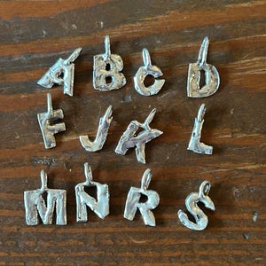 May include: A collection of silver-colored metal alphabet pendants, each with a loop for hanging. The letters A, B, C, D, F, J, K, L, M, N, R, and S are displayed. The pendants are on a wooden surface.