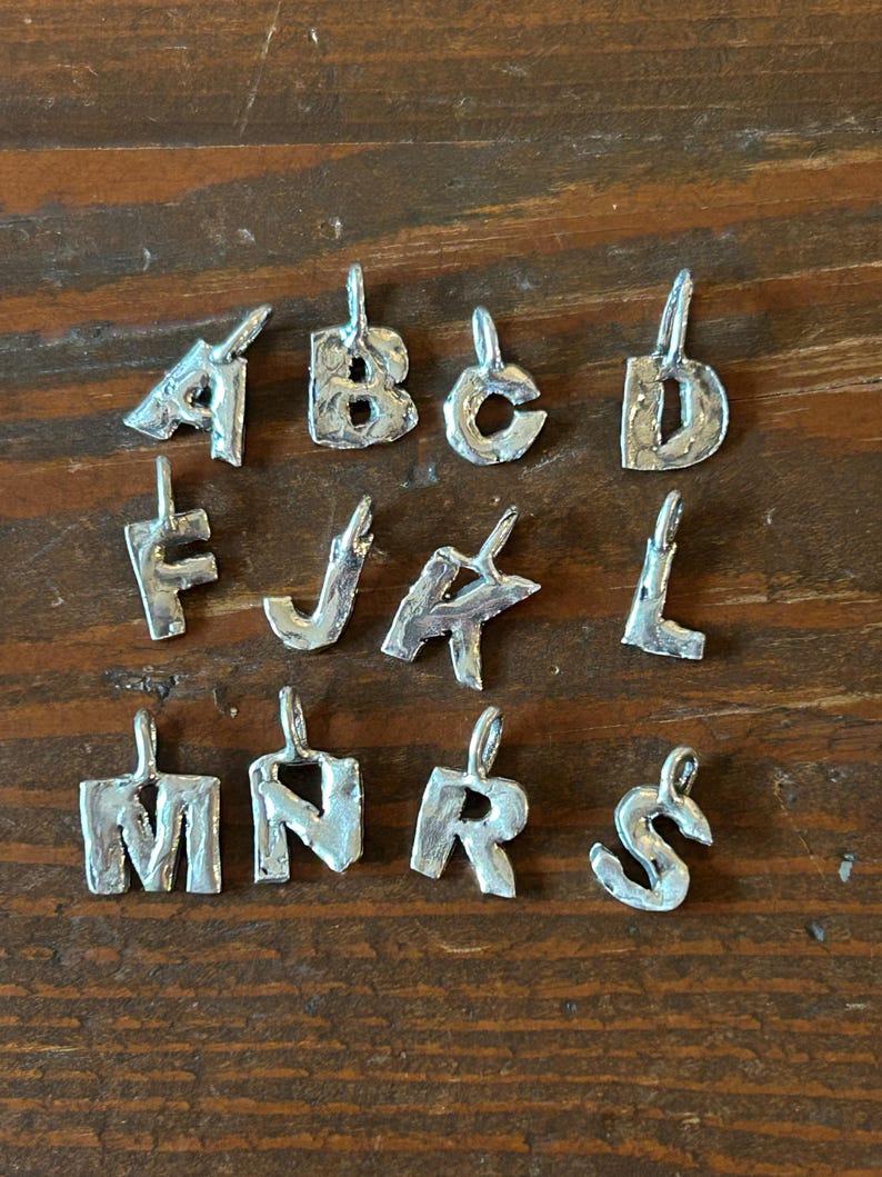 May include: A collection of silver-colored metal alphabet pendants, each with a loop for hanging. The letters A, B, C, D, F, J, K, L, M, N, R, and S are displayed. The pendants are on a wooden surface.