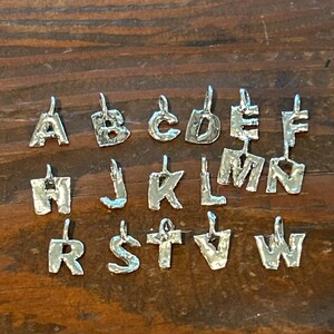 May include: Silver alphabet charms, each with a unique, textured design. The letters A through W are displayed, each with a small loop at the top for attaching to a chain or bracelet. The charms are arranged on a dark wooden surface.