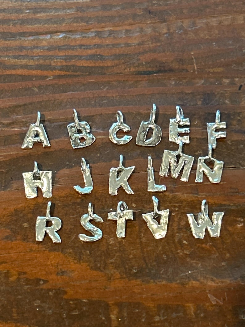May include: Silver alphabet charms, each with a unique, textured design. The letters A through W are displayed, each with a small loop at the top for attaching to a chain or bracelet. The charms are arranged on a dark wooden surface.
