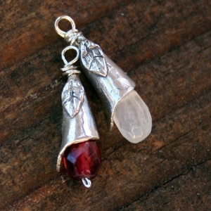 May include: Two silver-toned pendants with a leaf design. One pendant has a faceted, deep red gemstone, and the other has a clear, elongated gemstone. Both pendants have a loop at the top for hanging.