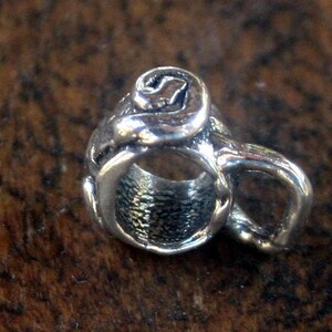 May include: A silver charm in the shape of a coffee cup with a swirl design on the top.