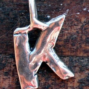 May include: A silver-colored pendant in the shape of the letter "K". The pendant has a textured, uneven surface and a loop at the top for a chain. The background is a dark, wooden surface.