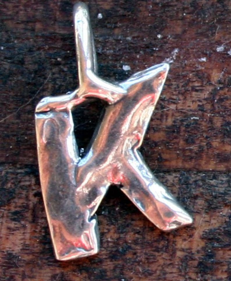 May include: A silver-colored pendant in the shape of the letter "K". The pendant has a textured, uneven surface and a loop at the top for a chain. The background is a dark, wooden surface.