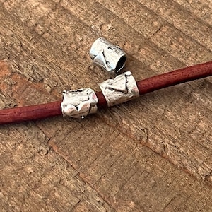 May include: Three silver-colored, textured metal beads. Two beads are strung on a maroon leather cord. The beads have a rough, irregular surface. The background is a wooden surface with visible grain.