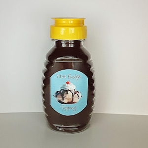 May include: A clear plastic bottle with a yellow lid filled with dark brown hot fudge topping. The label on the bottle says "Hot Fudge Topping" and features a cartoon image of a sundae with whipped cream, a cherry, and chocolate sauce.