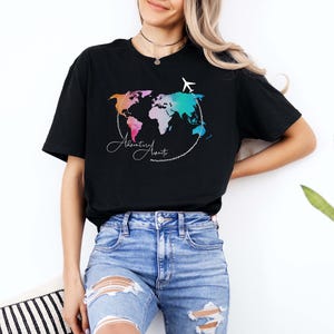 May include: Black t-shirt featuring a colorful world map design with the words "Adventure Awaits" in a script font. A white airplane graphic is above the map. The shirt is paired with blue jeans.