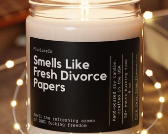 Puede incluir: Una vela de soja encendida en un tarro de cristal con una etiqueta negra que dice "Smells Like Fresh Divorce Papers". La vela está sobre un posavasos de madera, rodeada de luces de cadena. Una guitarra y un libro están en el fondo.