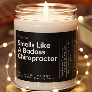 May include: A lit soy candle in a glass jar with a black label that reads "Smells Like A Badass Chiropractor." The candle sits on a small wooden coaster. The background includes a guitar and string lights.