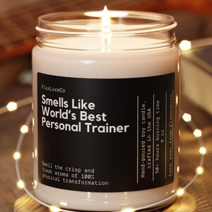 May include: A lit, hand-poured soy candle in a glass jar, labeled "Smells Like World's Best Personal Trainer." The candle sits on a wooden coaster, surrounded by string lights. The label also states the candle is crafted in the USA and has a 50+ hour burn time.