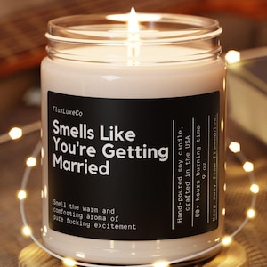 May include: A lit soy candle in a glass jar with a black label that reads "Smells Like You're Getting Married." The candle sits on a wooden coaster, surrounded by fairy lights. The candle is 255 grams.
