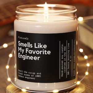 Puede incluir: Una vela de soja encendida en un tarro de cristal con una etiqueta negra que dice "Smells Like My Favorite Engineer". La vela está sobre un posavasos redondo de madera, rodeada de luces de cadena. Hecho a mano en EE. UU.