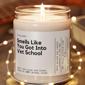 May include: A lit soy candle in a glass jar with the text "Smells Like You Got Into Vet School." The candle sits on a wooden coaster, surrounded by string lights. The candle is hand-poured in the USA and has a 50+ hour burn time.
