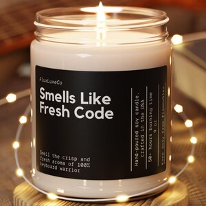 Op de afbeelding: Een brandende sojakaars in een glazen pot met een zwart etiket met de tekst "Smells Like Fresh Code". De kaars staat op een houten onderzetter, omringd door lichtslingers. Op de achtergrond een gitaar en een boek.