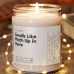 May include: A lit soy candle in a clear glass jar with the text "Smells Like Math Up In Here." The candle sits on a wooden coaster, surrounded by string lights. The background includes a guitar and a book.