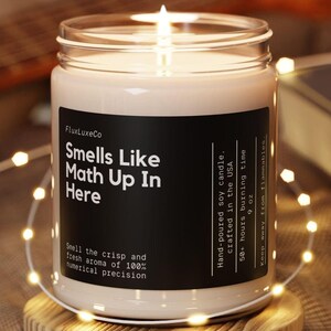 May include: A lit soy candle in a glass jar with a black label that reads "Smells Like Math Up In Here." The candle sits on a wooden coaster, surrounded by string lights. The text on the label also says "Hand-poured soy candle, crafted in the USA, 9 oz, 50+ hours burning time."