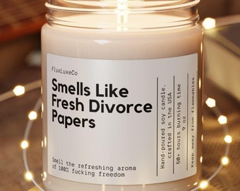Puede incluir: Una vela de soja encendida en un tarro de cristal con el texto "Smells Like Fresh Divorce Papers". La vela está sobre un posavasos de bambú, rodeada de luces de cadena. La etiqueta también dice "Smell the refreshing aroma of 100% fucking freedom."