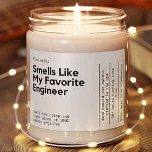 Pode incluir: Uma vela de soja acesa num frasco de vidro transparente, com a etiqueta "Smells Like My Favorite Engineer". A vela está sobre um descanso de madeira, rodeada por luzes de corda. A vela pesa 255 g e tem um tempo de queima de 50+ horas.