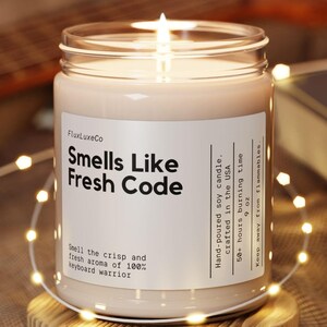 Puede incluir: Una vela de soja encendida en un tarro de cristal con el texto "Smells Like Fresh Code". La vela está sobre un posavasos de madera, rodeada de luces de cadena. La etiqueta indica que está vertida a mano en EE. UU. y tiene un tiempo de combustión de 50+ horas.