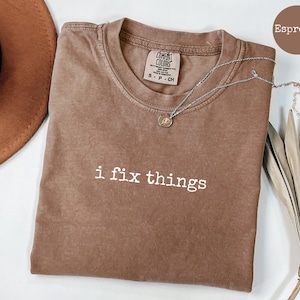 May include: Espresso-colored t-shirt with the phrase "i fix things" in white. The shirt has a crew neck and short sleeves. A silver necklace with a pendant is draped across the shirt.