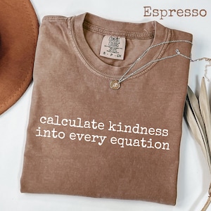 Può includere: T-shirt color espresso con la frase bianca "calculate kindness into every equation". Una collana d'argento con un piccolo ciondolo è appoggiata sulla maglietta. La maglietta è del marchio Comfort Colors.