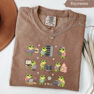 May include: An espresso-colored t-shirt featuring a graphic print of nine green frogs performing various IT and computer tasks, including working on servers, using laptops, and managing network equipment. The shirt has a crew neck and a Comfort Colors label.