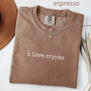 May include: Espresso-coloured t-shirt with the text "i love crypto" in white. The shirt has a crew neck and a Comfort Colors tag. A silver necklace with a pendant is draped across the shirt.